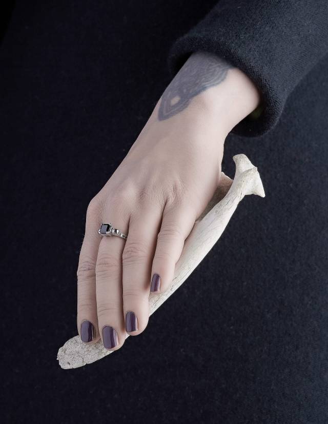 Thyone ist ein gothic Damenring mit Totenköpfen und schwarzen Diamanten. Eine stabile Fassung hält den viereckigen, quadratisch geschliffenen Edelstein in der Mitte. Seitlich unter den Totenköpfen ist der Ring mit je drei Diamanten besetzt. Gezeigt wird der Ring an einer Frauenhand.