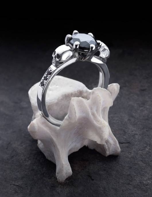 Thana ist ein mystischer Ring mit Totenköpfen und funkelnden Diamanten. Eine zarte Fassung hält den runden, facettierten Edelstein in der Mitte. An den Seiten ist der Ring mit je drei Diamanten besetzt. Gezeigt wird der Ring auf einem Knochen stehend.