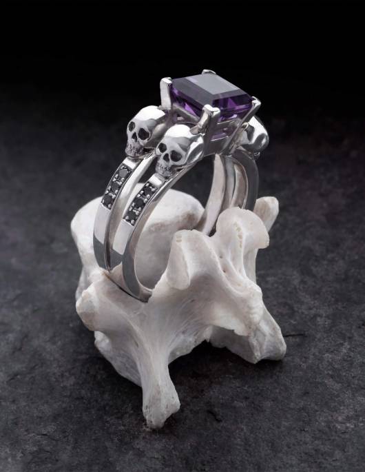 Lethe ist ein massiver Totenkopf Damenring mit schwarzen Diamanten und einem eckigen Amethyst Edelstein aus der Goldschmiede Kipkalinka. Gezeigt auf einem Knochen.