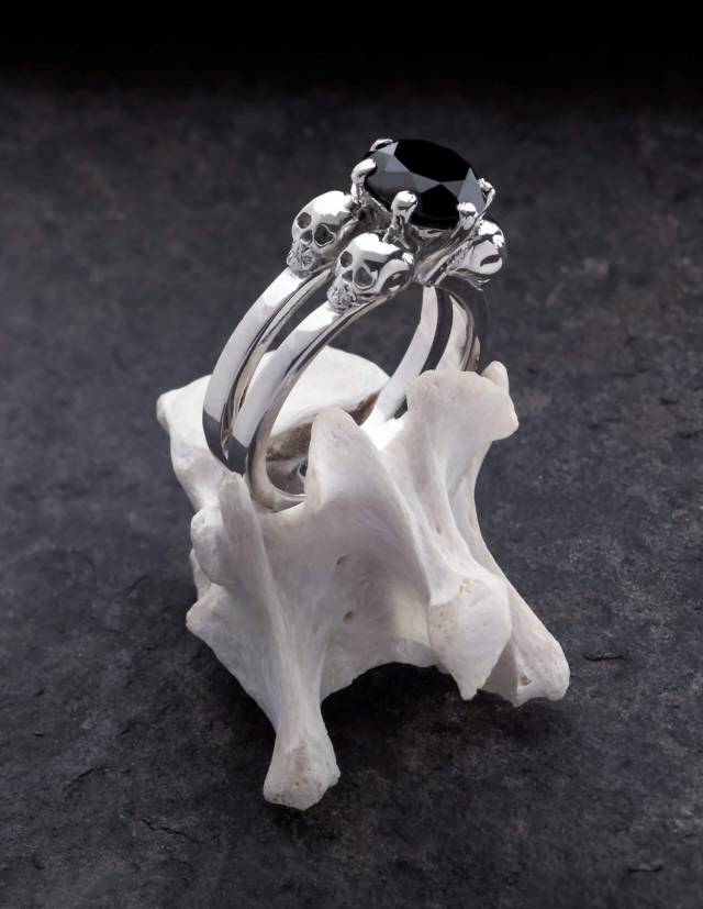 Lilith ist ein edler Totenkopfring für gothic Frauen. Der schlichte Ring ist aus Silber gefertigt und trägt zwischen vier kleinen Totenköpfen einen schwarzen Edelstein in der Mitte. Gezeigt auf einem Knochen stehend.