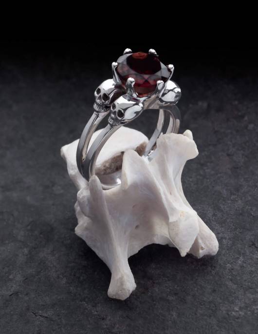 Varla es un anillo de compromiso con pequeñas calaveras de plata. Entre las cuatro calaveras hay una gran gema redonda. El anillo está colocado sobre un hueso.