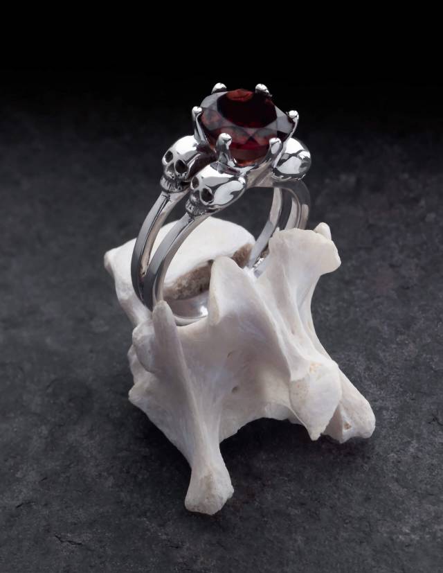 Varla ist ein Verlobungsring mit kleinen Totenköpfen aus Silber. Zwischen den vier Totenschädeln sitzt ein großer, runder Edelstein. Der Ring ist auf einem Knochen drapiert.