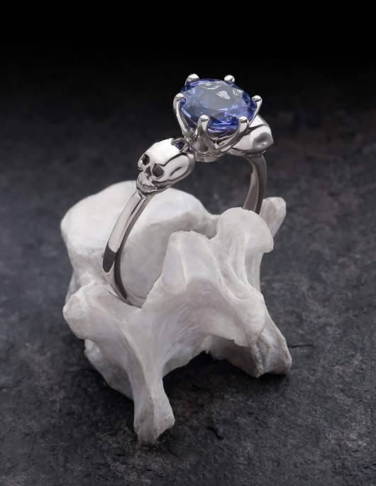 Wanda est une élégante bague tête de mort avec une grande pierre précieuse à facettes. Les deux têtes de mort détaillées tiennent la monture en leur centre. La bague sur la photo est en or blanc avec une tanzanite et est présentée sur un os.