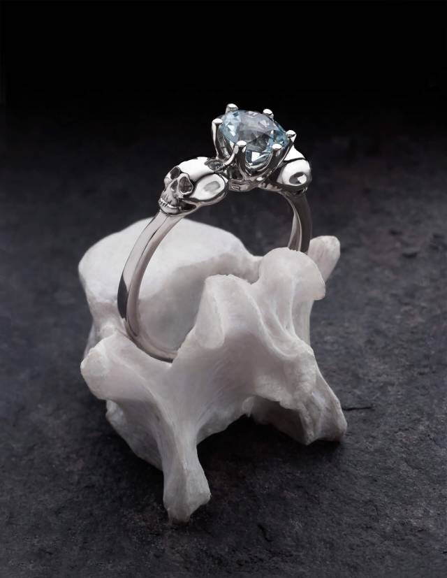 WANDIKA es un delicado anillo de oro blanco con calaveras. Lleva una gema de aguamarina azul claro en su centro. Se muestra en un hueso.