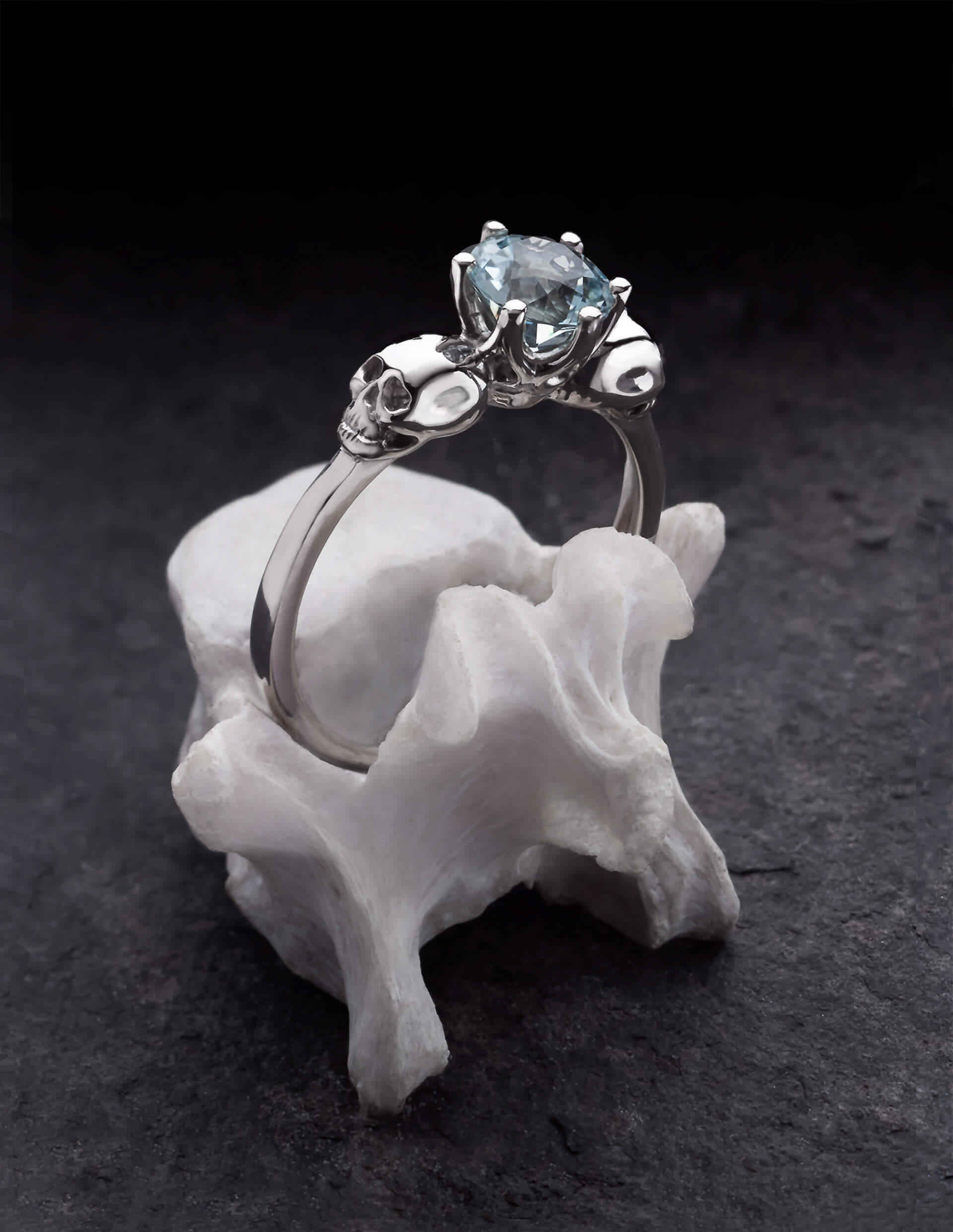 WANDIKA ist ein zierlicher Weißgold Ring mit Totenköpfen. Er trägt einen hellblauen Aquamarin Edelstein in seiner Mitte. Gezeigt auf einem Knochen.