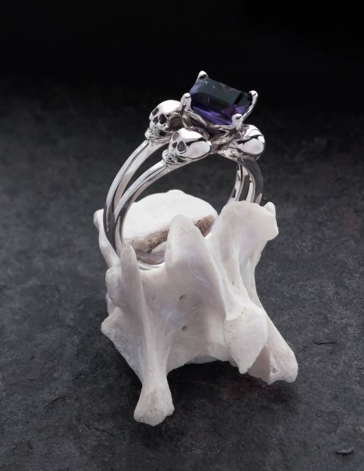 Zorya es un anillo de calavera de plata con una piedra angular para mujer. La amatista se sitúa en el centro entre cuatro pequeñas calaveras. Se muestra en un hueso.