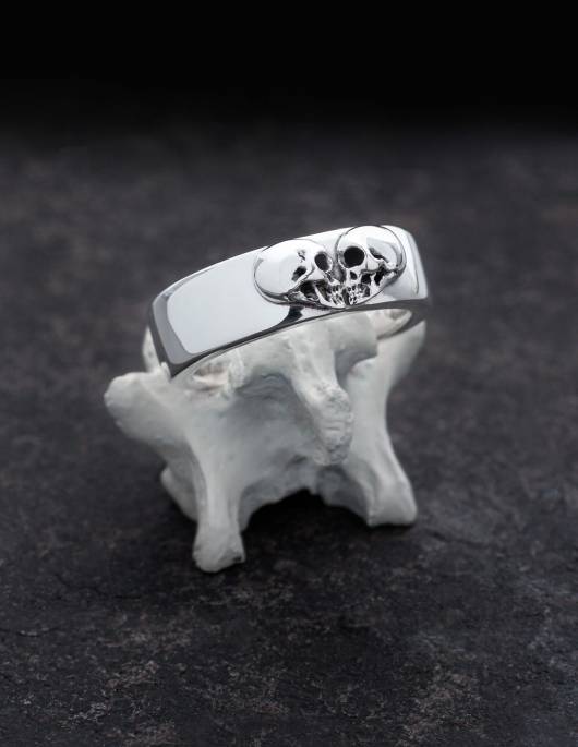 Kissing Skulls est une bague gothique massive en argent. Deux têtes de mort de profil sont disposées en forme de cœur. La forme de l'anneau est large et légèrement bombée à plat avec des bords arrondis. L'anneau est présenté sur un os.