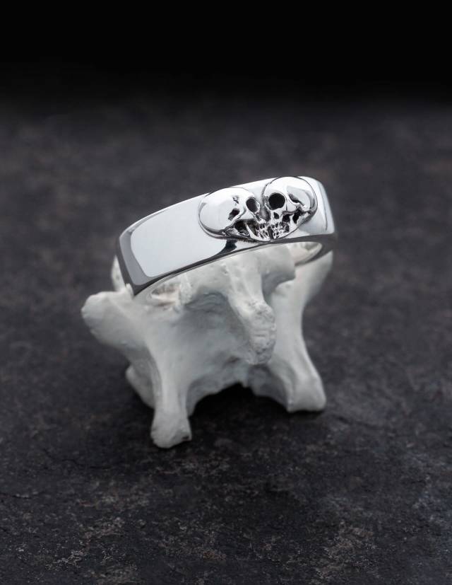 Kissing Skulls is a solid gothic silver ring. Two skulls in profile are arranged in the shape of a heart. The shape of the ring is wide and slightly flat domed with rounded edges. The ring is shown on a bone.