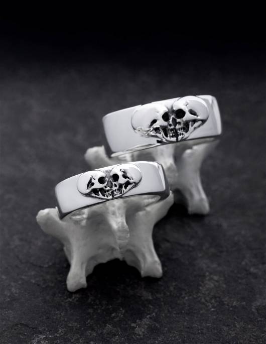 Alt tag (image title as continuous text): Kissing Skulls are a pair of heavy 925 silver wedding rings for real rockers. The two skulls in profile form a heart shape. The wedding rings are wide and flat domed. The rings can be seen on bones.