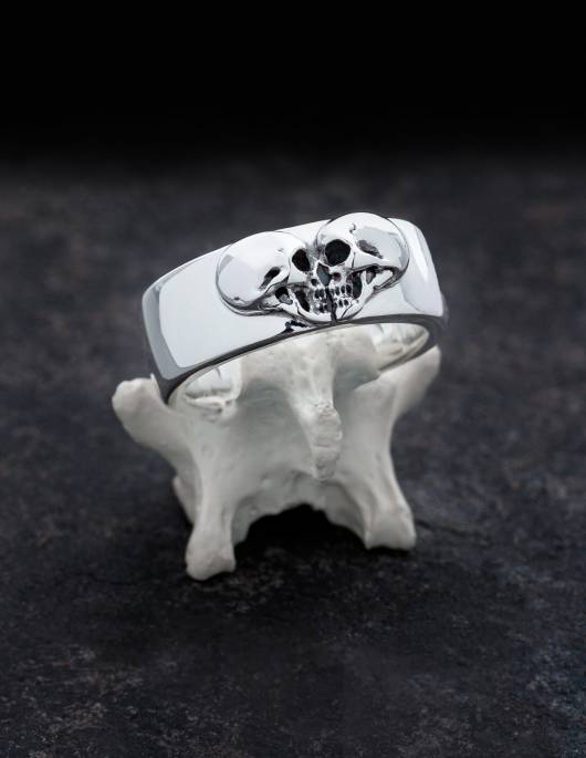 Kissing Skulls è una fede nuziale gotica pesante. Due teschi di profilo sono disposti a forma di cuore. La forma dell'anello è ampia e leggermente piatta a cupola con bordi arrotondati. In questa foto l'anello è drappeggiato su un osso.