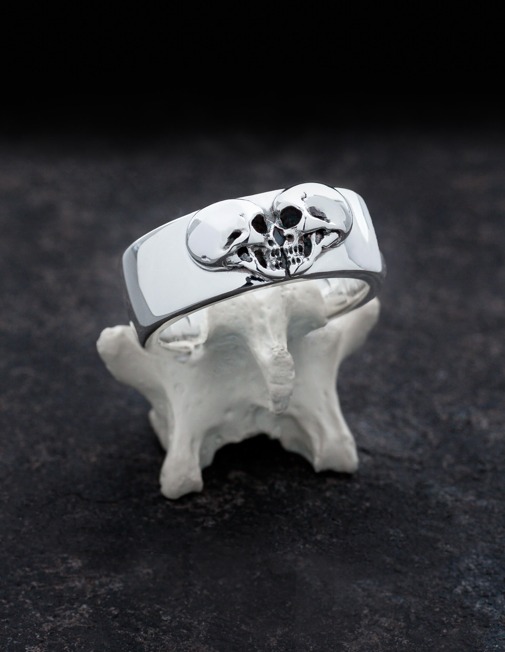 Kissing Skulls ist ein schwerer gothic Ehering. Zwei Totenköpfe im Profil sind in Form eines Herzens angeordnet. Die Form des Ringes ist breit und leicht flach gewölbt mit abgerundeten Kanten. Der Ring ist in diesem Bild auf einem Knochen drapiert.