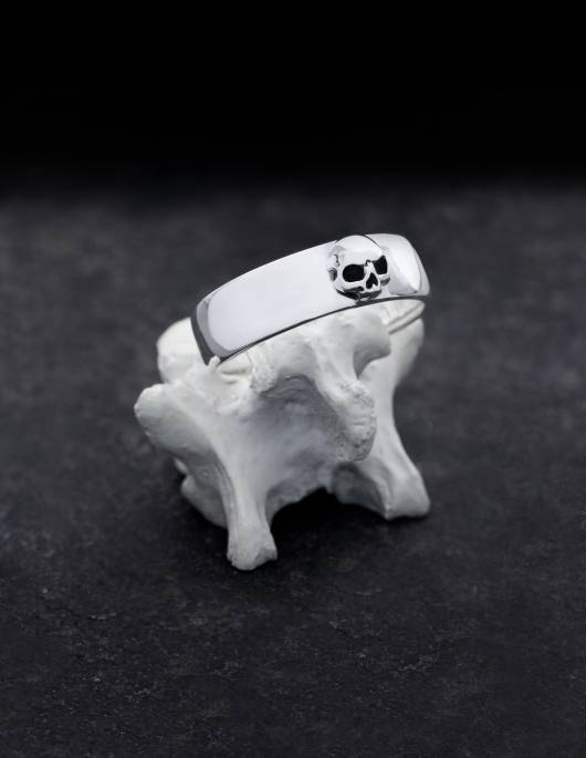 Amunet is a discreet band ring made of silver with a sculpted skull without a lower jaw. The ring has a flat shape, slightly curved on the inside and outside, the edges are rounded. It is presented standing on a bone.