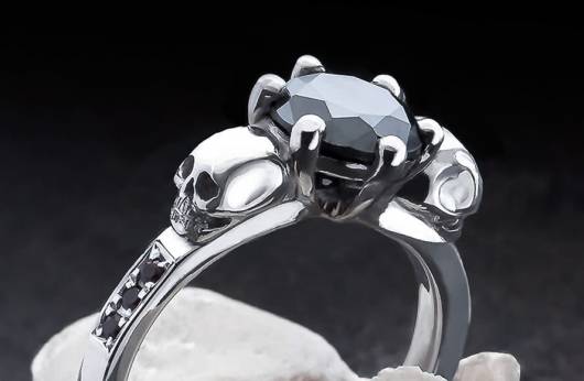 Auf dem Bild ist ein zarter Ring mit kleinen Totenköpfen und geschliffenen Edelsteinen zu sehen. Der große Stein in der Mitte ist ein schwarzer Edelstein, die kleinen an den Seiten schwarze Diamanten. Das Bild führt zur Kategorie "Totenkopf-Ringe mit Edelsteinen" im Shop von Kipkalinka.