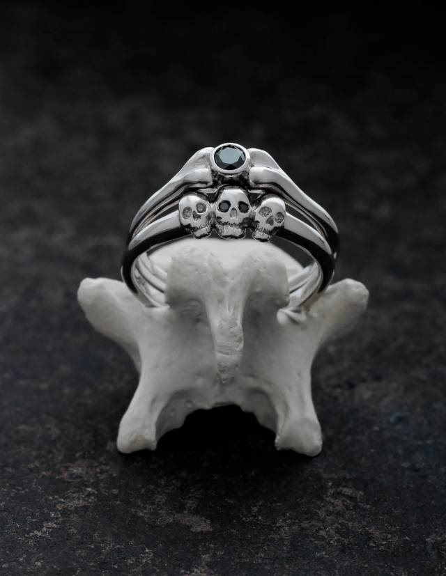 Two small rings are presented standing on a bone.... One has the shape of a bone and a round gemstone. The other three tiny skulls. The rings are made of 925 silver.