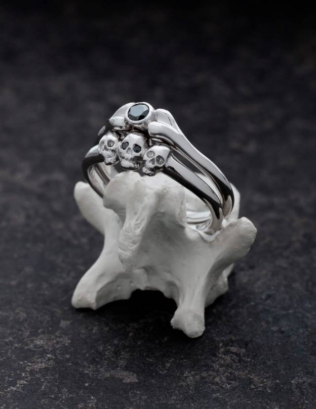 Two small silver rings on a bone. One has the shape of a bone and a round gemstone. The other is decorated with three tiny skulls.