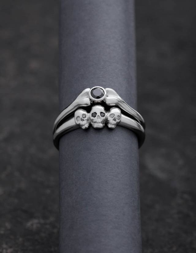 Dos anillos pequeños. Uno tiene la forma de un hueso y una piedra preciosa redonda. El otro tiene tres cráneos pequeños. Los anillos son de plata 925.