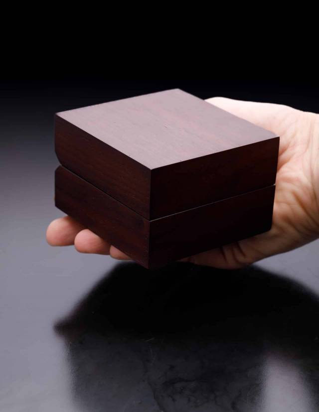 Ring box with walnut veneer lies closed on one hand against dark background