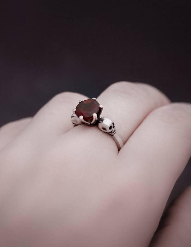 A dainty skull ring with two skulls and a red gemstone worn on a hand.