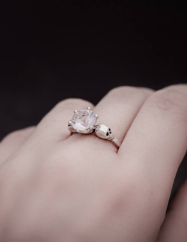 A tiny skull engagement ring with light pink stone shown on a hand. THe ring is named Wanda.