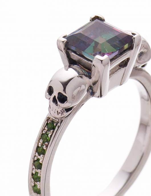 A detailed skull engagement ring made of silver with square mystic topaz gemstone and green stones down the bands.