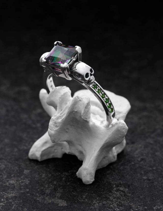 Ein silberner Totenkopfring mit grünen Edelsteinen auf einem Knochen präsentiert, Modell THYONE