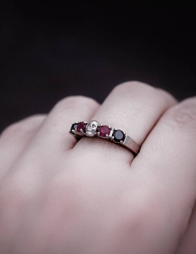 Close up of a dainty skull ring made of solid white gold with rubys and black diamonds shown on a hand.